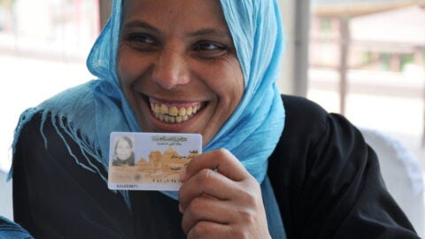 Woman with blue headscarf holding up national ID card and smiling.