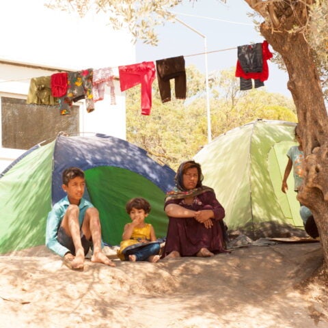 Woman and two children seated on the dirt in front of a green camping tent in a refugee camp. Laundry hanging to dry on laundry line above them.