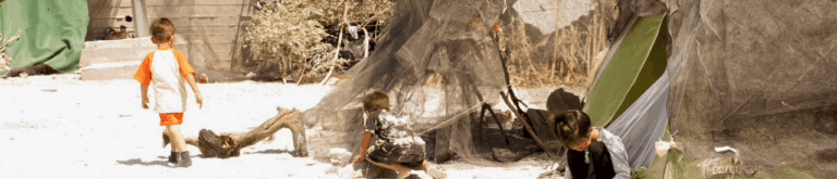 Children playing in a refugee camp, by a concrete wall and nets.