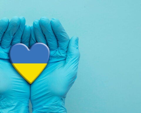 A pair of hands in blue surgical gloves cradles a hear in the colors of the Ukrainian flag against a light blue background.