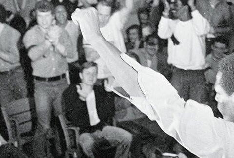 Man with back to camera raises left first in front of audience of standing and sitting people.