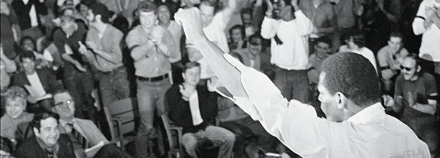 Man with back to camera raises left first in front of audience of standing and sitting people.