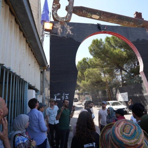 Group of people listening to man in green polo shirt under black and red arch on the street in Palestine