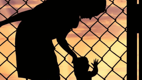 Image of black figure of woman bending over and holding hand of small child against barbed wire fence and sunset.