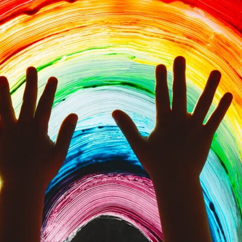 Close-up photo of child's hands touch painting rainbow on window.