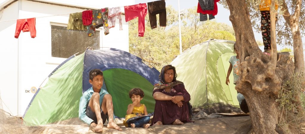 Woman and two children seated on the dirt in front of a green camping tent in a refugee camp. Laundry hanging to dry on laundry line above them.