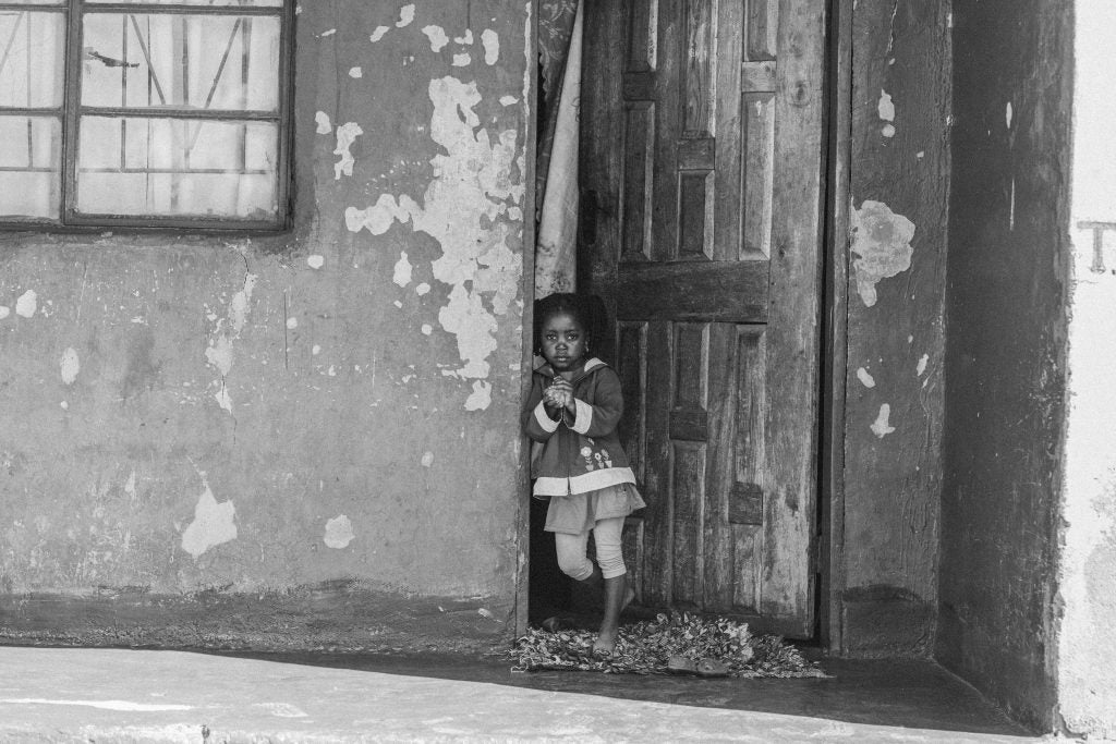 Black and white image of young girl standing in the doorway of a dilapidated house.