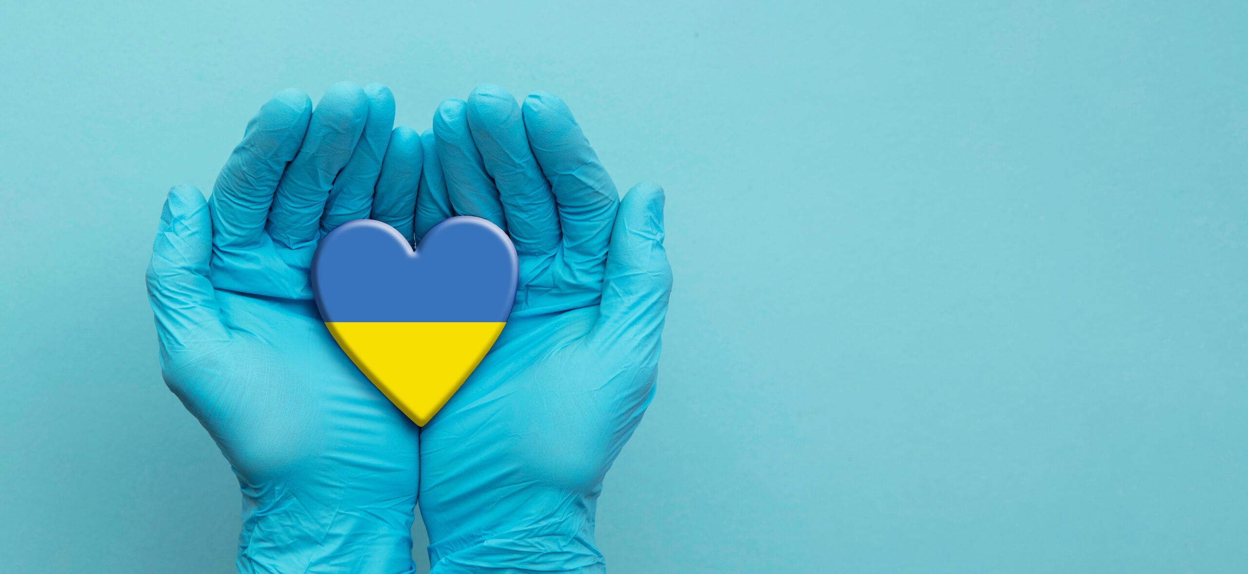 A pair of hands in blue surgical gloves cradles a hear in the colors of the Ukrainian flag against a light blue background.