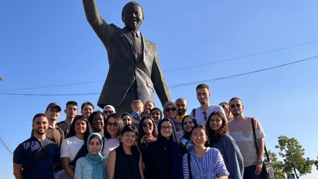 The 2023 cohort of the inaugural Palestine Social Medicine course standing in front of the statue of a man with a raised fist in Palestine.