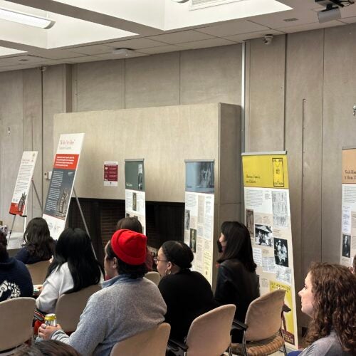 Seated audience listening to presentations of featured speakers during the opening of the Exhibition “We Are Not Alone”: Legacies of Eugenics at Harvard's Countway Library. Some of the exhibition material is visible between the audience and concrete wall.