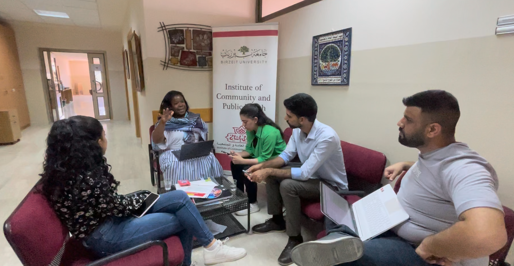 Seated woman speaking to group of four seated students in a hallway at Birzeit University Institute of Community and Public Health