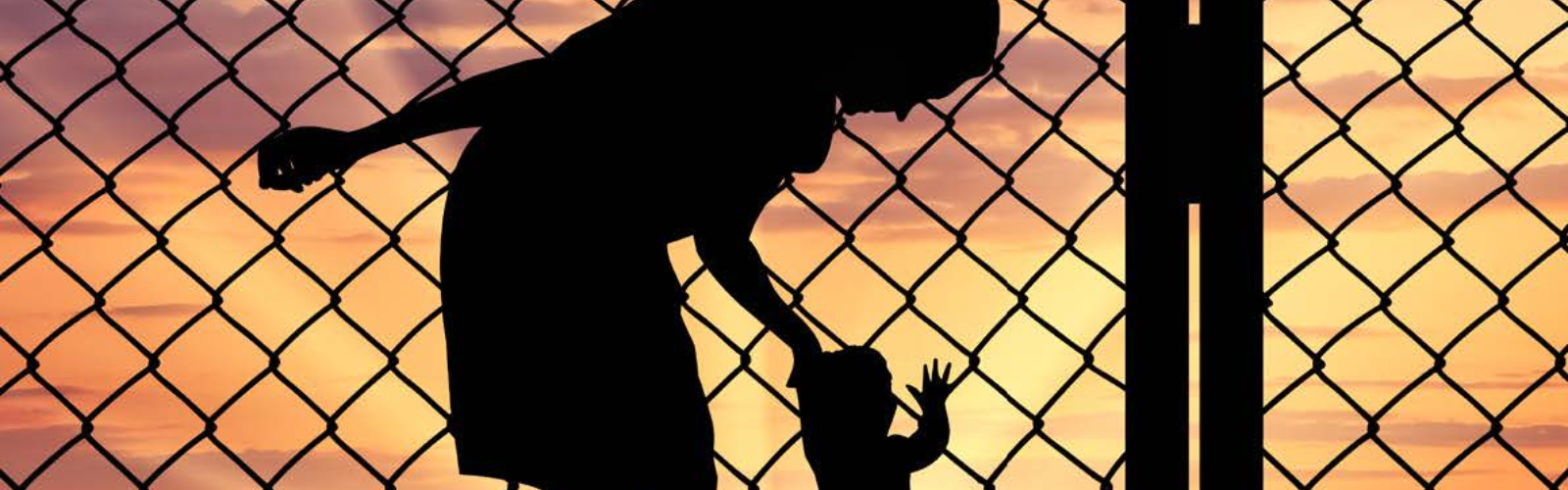 Image of black figure of woman bending over and holding hand of small child against barbed wire fence and sunset.