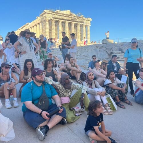 Members of 2024 cohort of intensive summer course on migration and refugee studies in Greece seated and standing on Acropolis Rock in Athens Greece, listening to presentation by tour guide. Parthenon in the background.