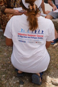 Person kneeling with back to camera wearing t-shirt that says intensive summer course on migration and refugee studies.