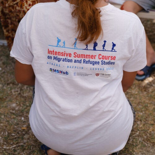 Person kneeling with back to camera wearing t-shirt that says intensive summer course on migration and refugee studies.